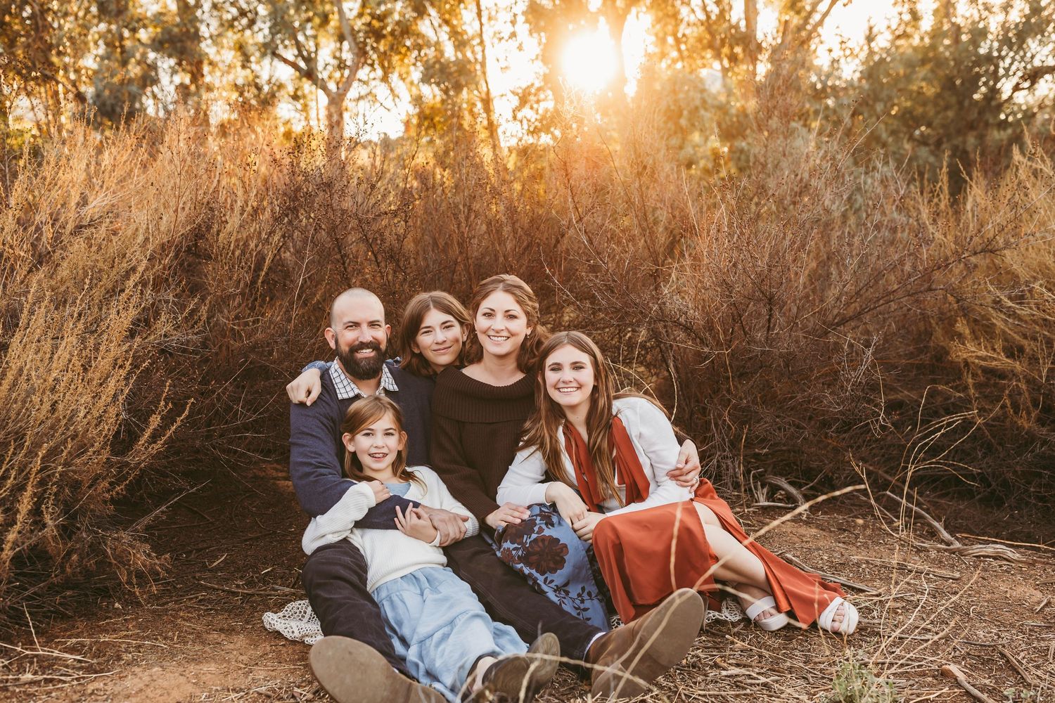 A group relaxes together on dry grass at sunset with golden light filtering through trees in the background.