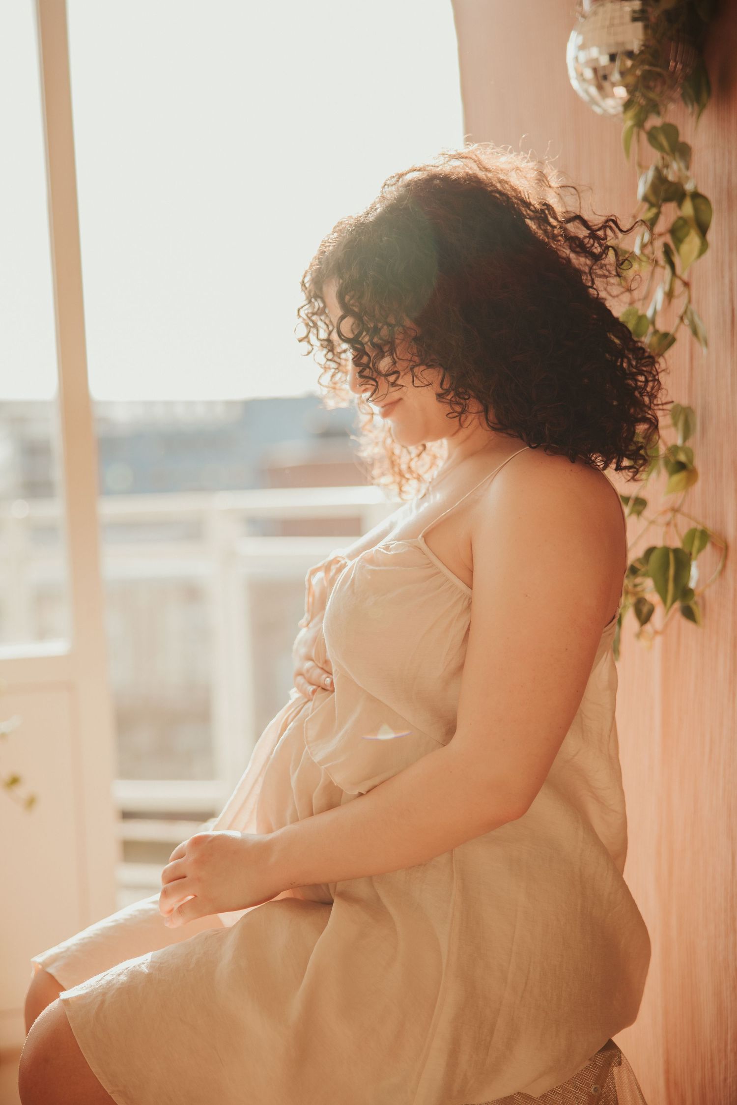 Sunlit silhouette of expecting mother with curly hair sitting by window with warm glowing light.