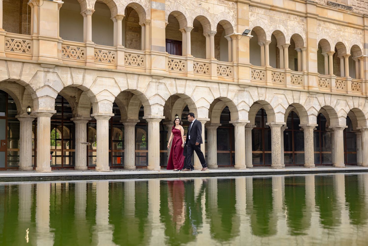 Couple in a red dress and dark suit walk hand in hand along an arcaded historic building, reflected in a calm pool of water.