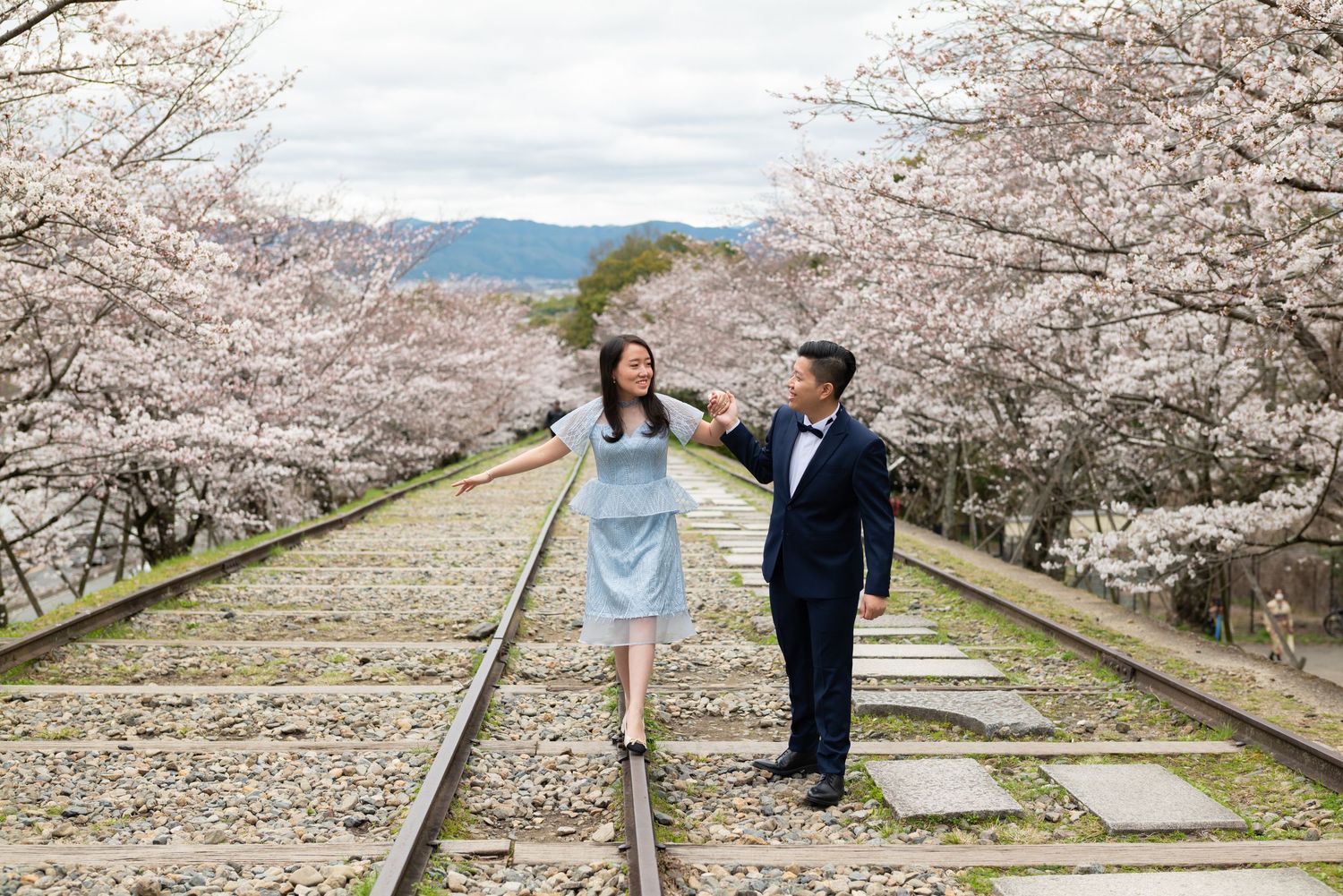 Couple holding hands while walking on old railroad tracks lined with blooming pink cherry blossom trees in spring.
