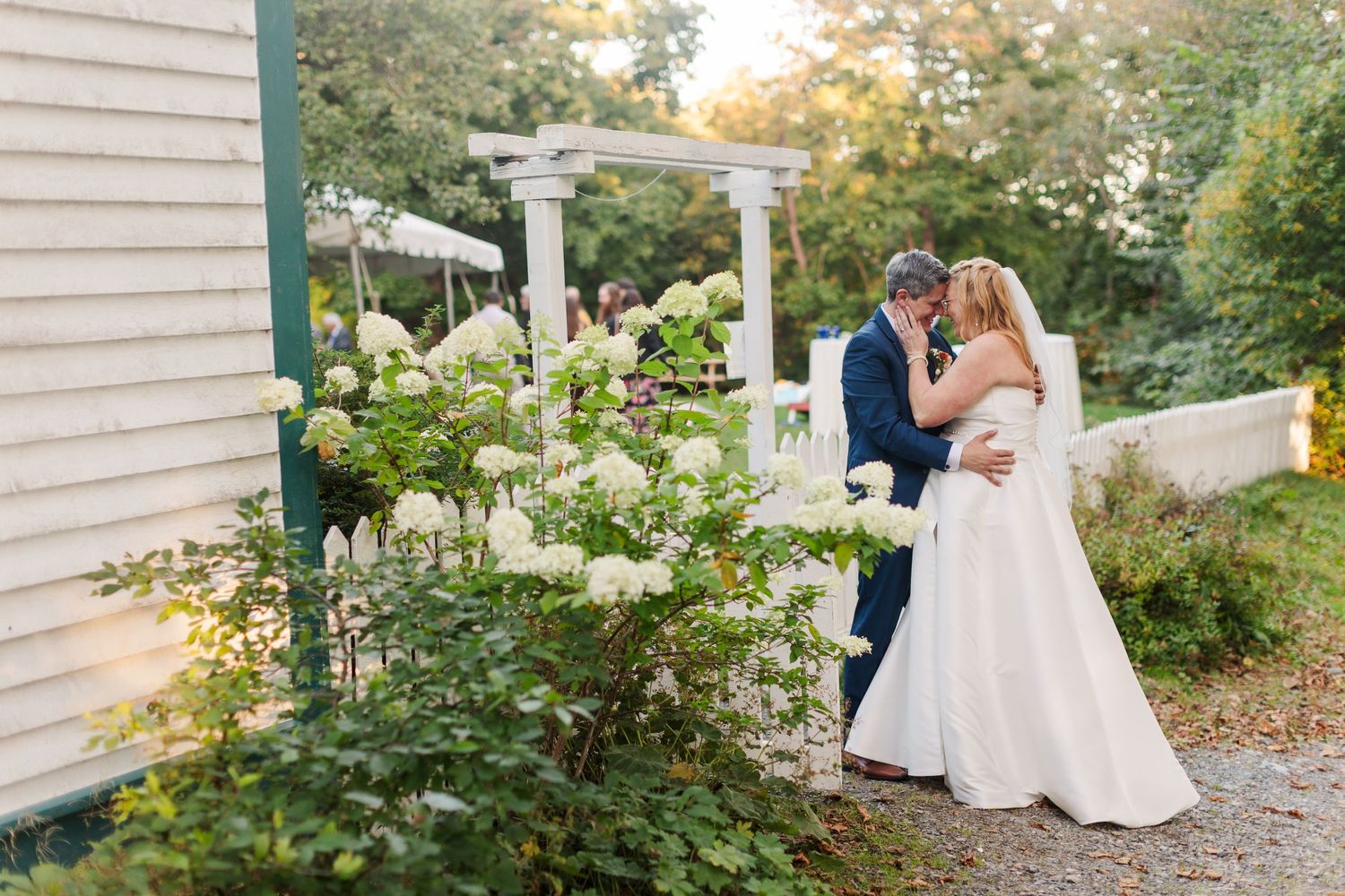 Bride in a white gown and groom in a dark suit embrace near blooming white hydrangeas beside a garden pergola at an outdoor wedding.