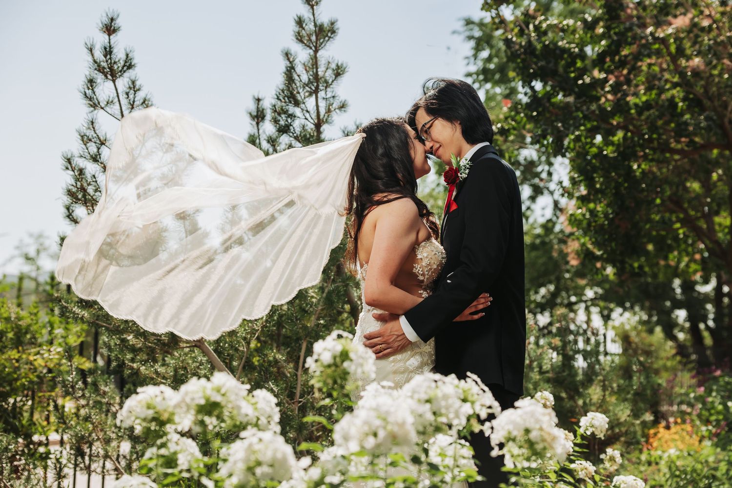Bride and groom kissing in garden with veil flowing in wind, surrounded by white hydrangeas and lush greenery.