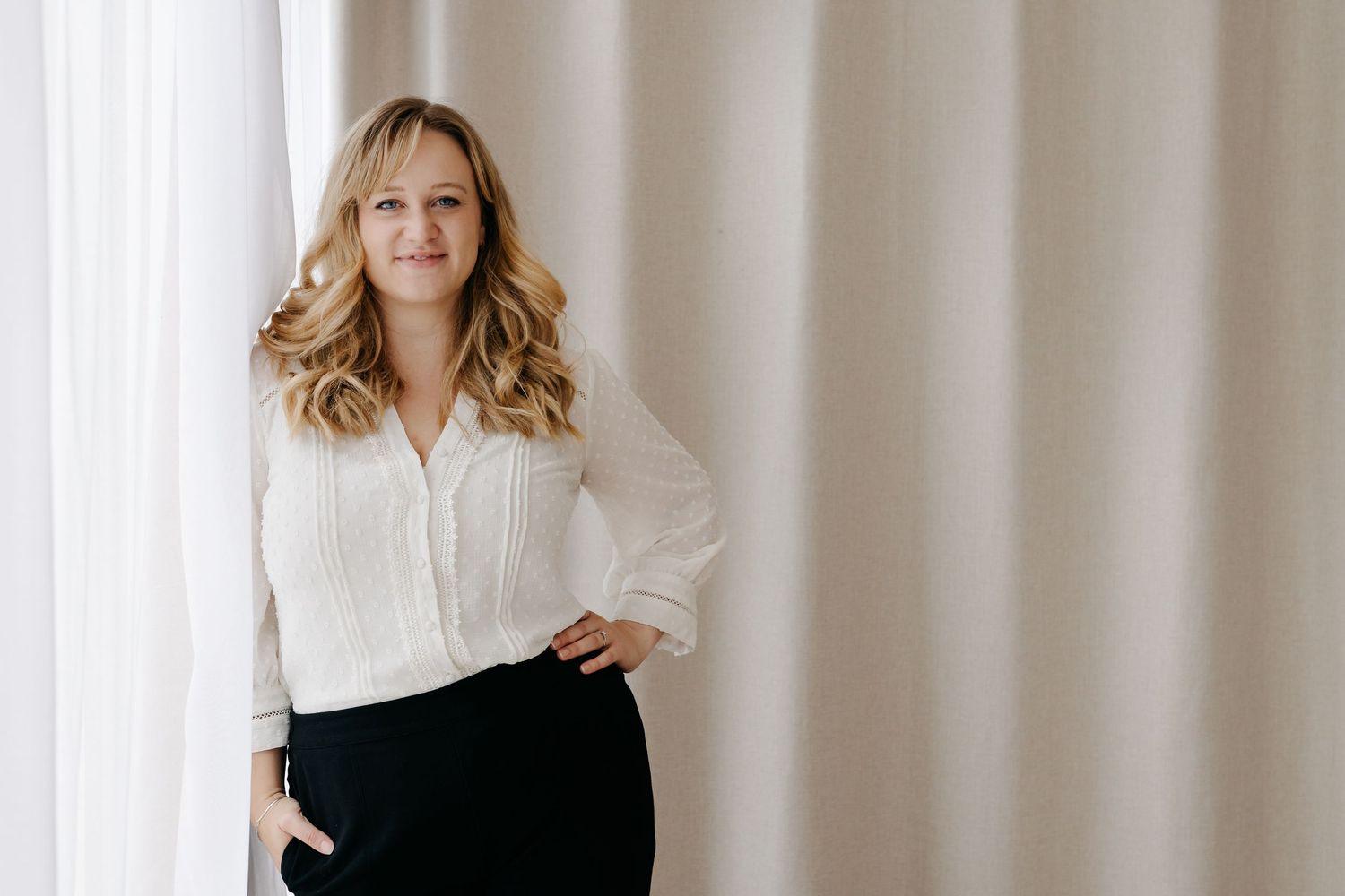 A confident blonde woman in a white blouse and black skirt poses gracefully beside sheer white curtains. Studio Headshot Session