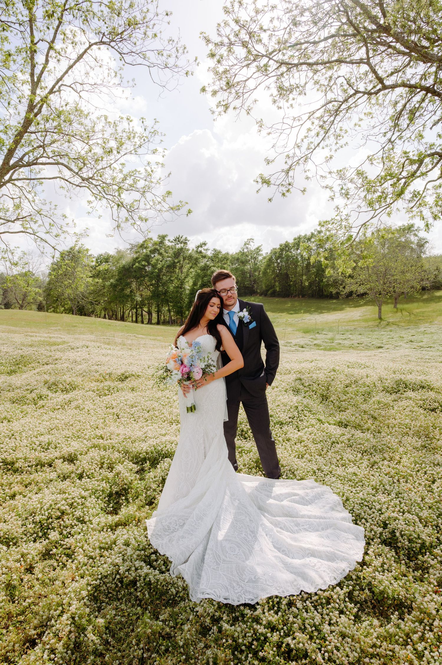 Bride and groom embrace in a lush green field surrounded by trees on their wedding day.