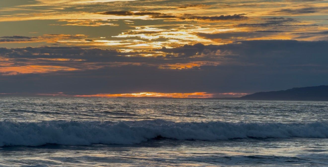 Golden sunset over ocean waves with dramatic cloudy sky reflecting warm hues of orange and blue at the beach.