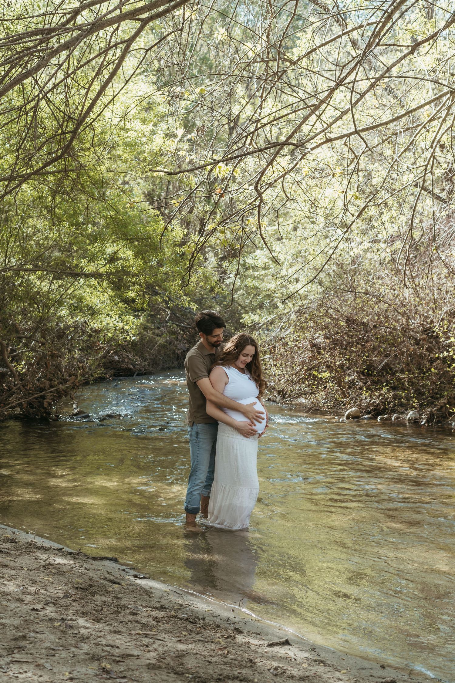 Couple embracing on rocky beach at golden sunset, pregnant woman in floral dress with partner in black shirt and jeans.
