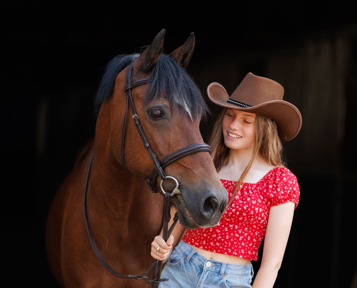 A person leads a palomino horse along a sunlit forest trail surrounded by tall evergreen trees.