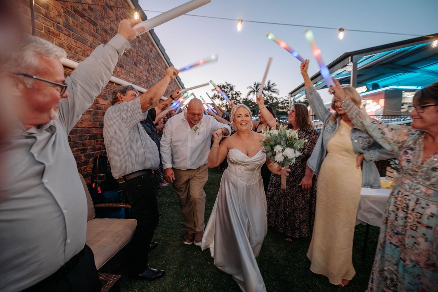 Bride walks through cheering guests waving glow sticks at an outdoor evening wedding reception.