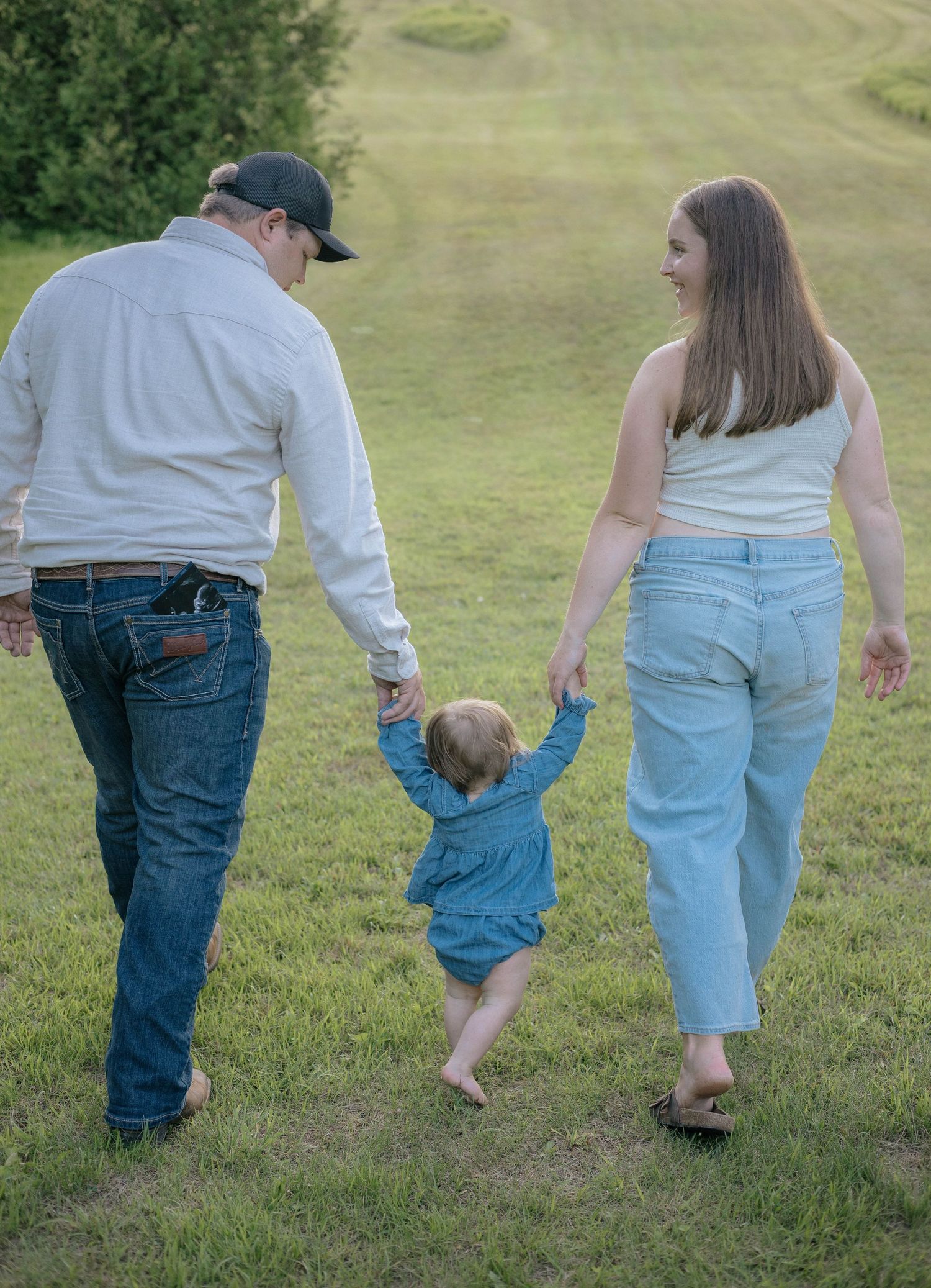 A young family walks hand-in-hand across a green grassy field, parents guiding their toddler in a denim outfit.
