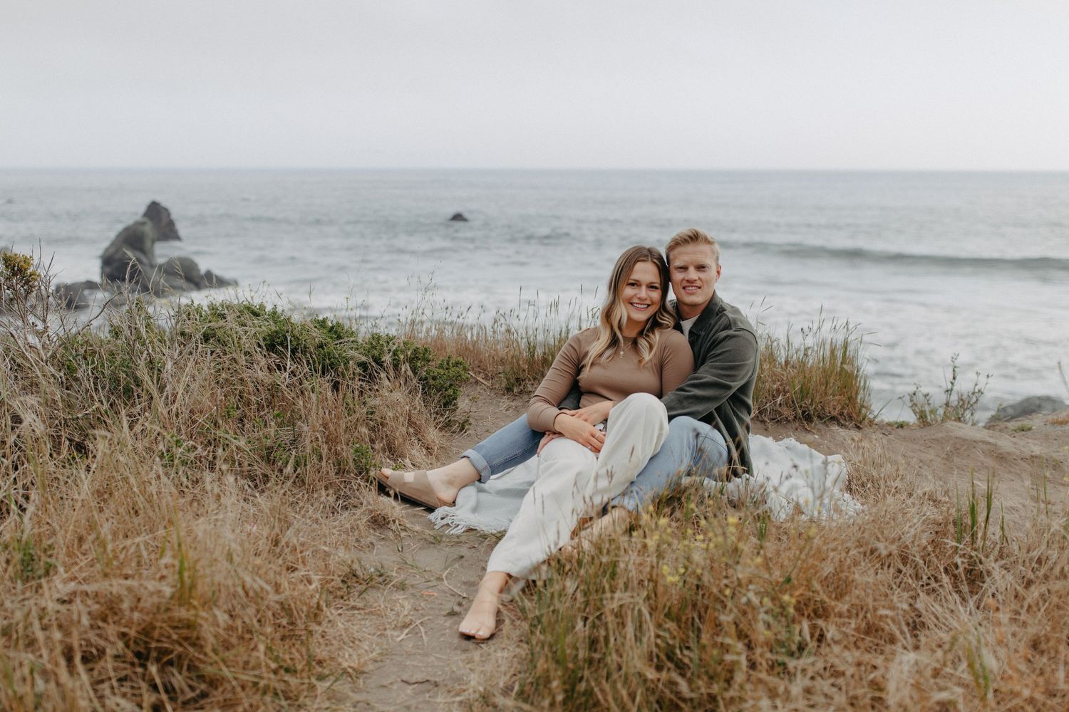 A couple sits together on coastal cliffs overlooking the ocean on a cloudy day.