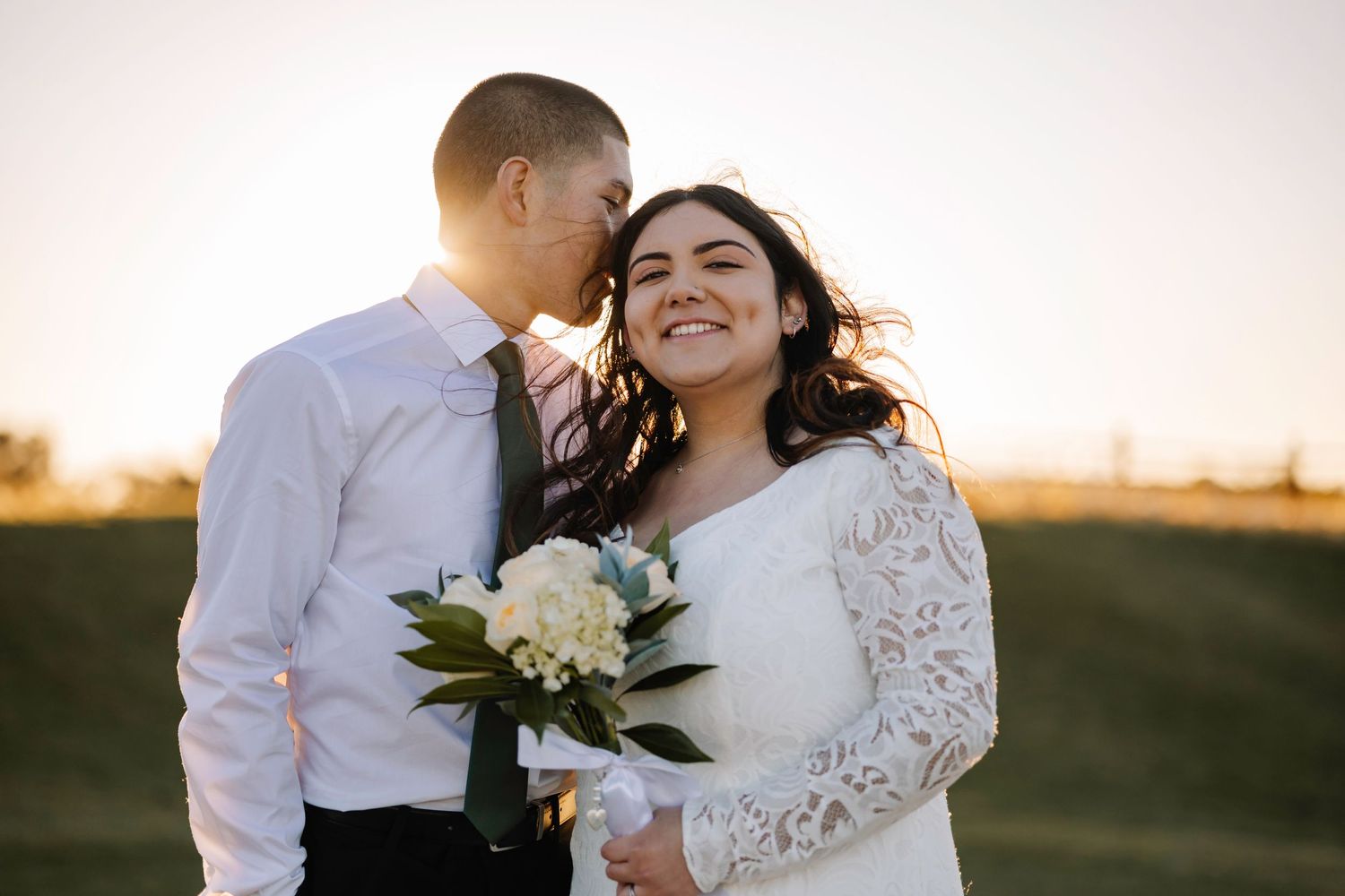 A wedding couple shares an intimate moment at sunset with white flowers in the bride's hands.