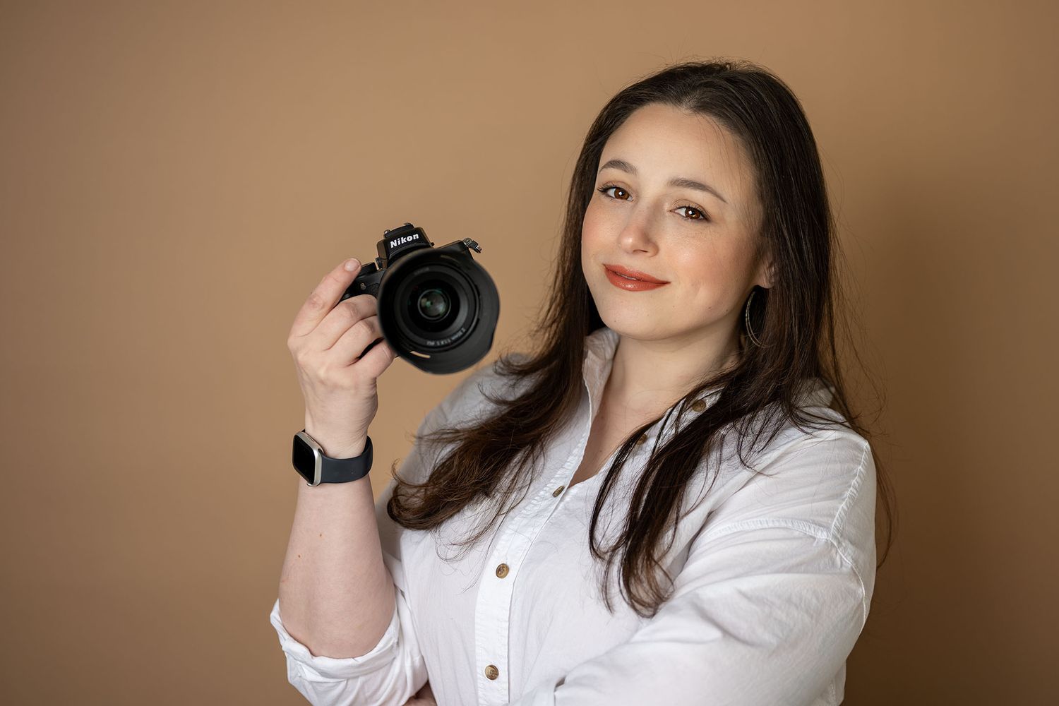 Smiling woman in white shirt holding a DSLR camera against beige background.