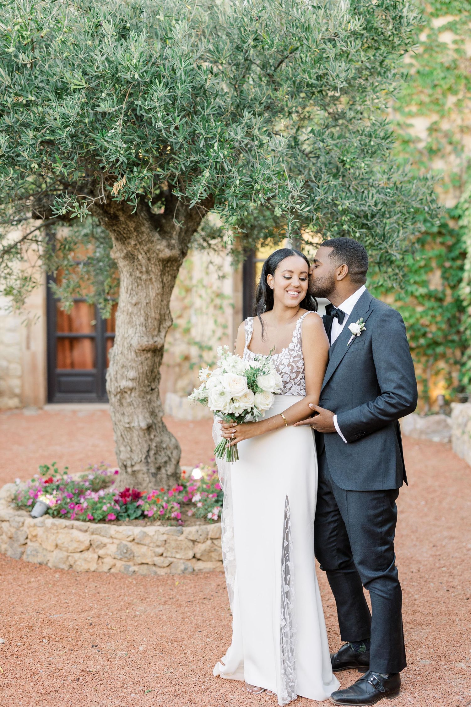 Wedding couple shares intimate moment under olive tree in Mediterranean garden setting.