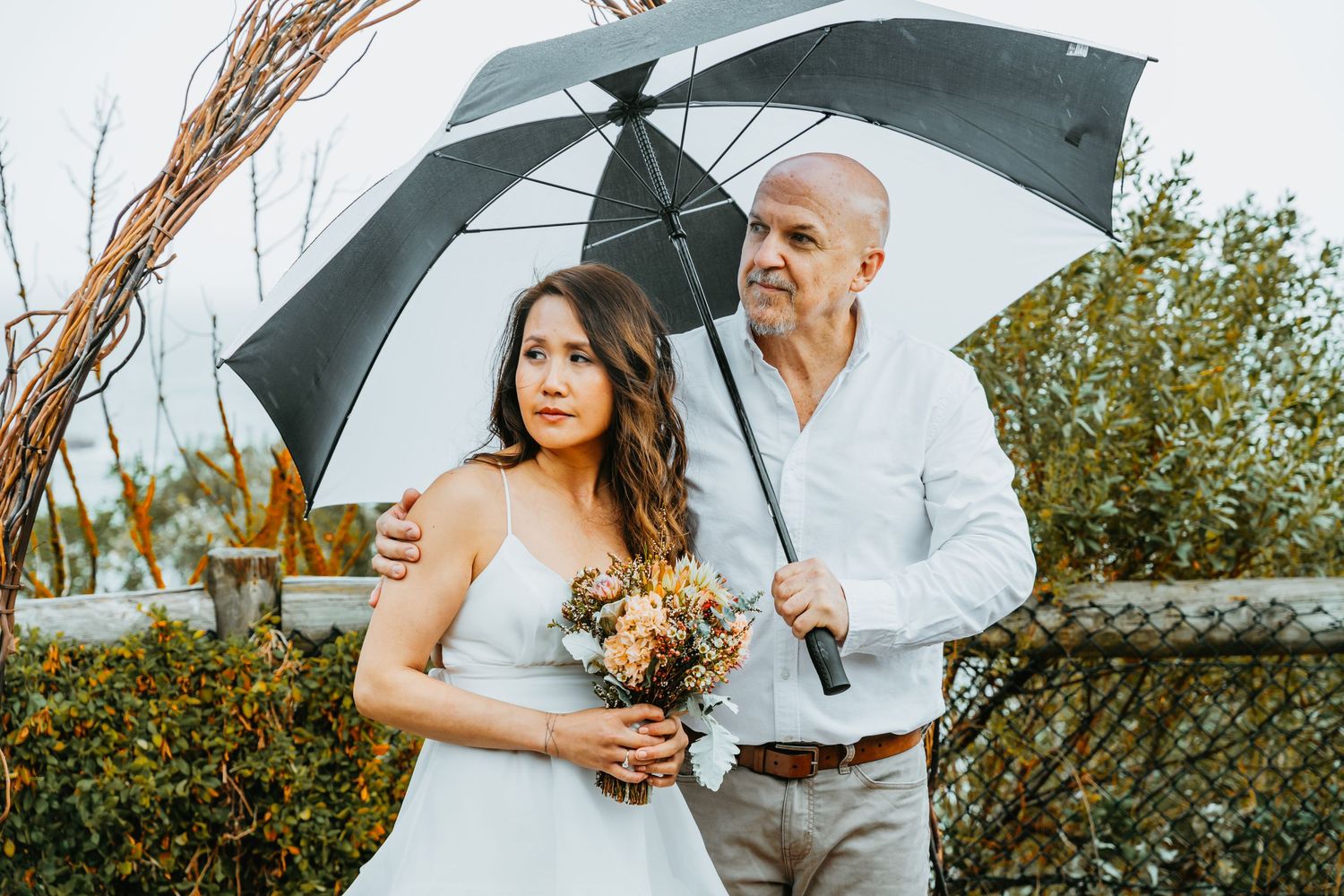 A couple stands together under a white umbrella during a wedding photoshoot in an outdoor garden setting.