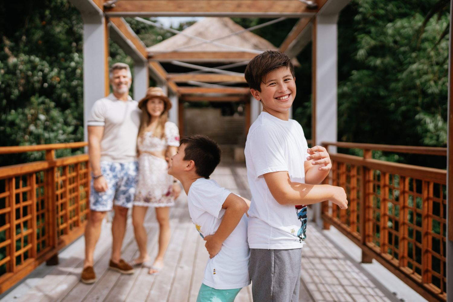 Family in white clothing poses together on wooden bridge walkway surrounded by trees.