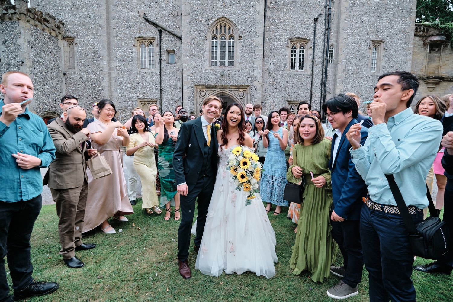 Wedding party gathered outside stone church celebrating with guests in joyful candid moment at Castle Goring