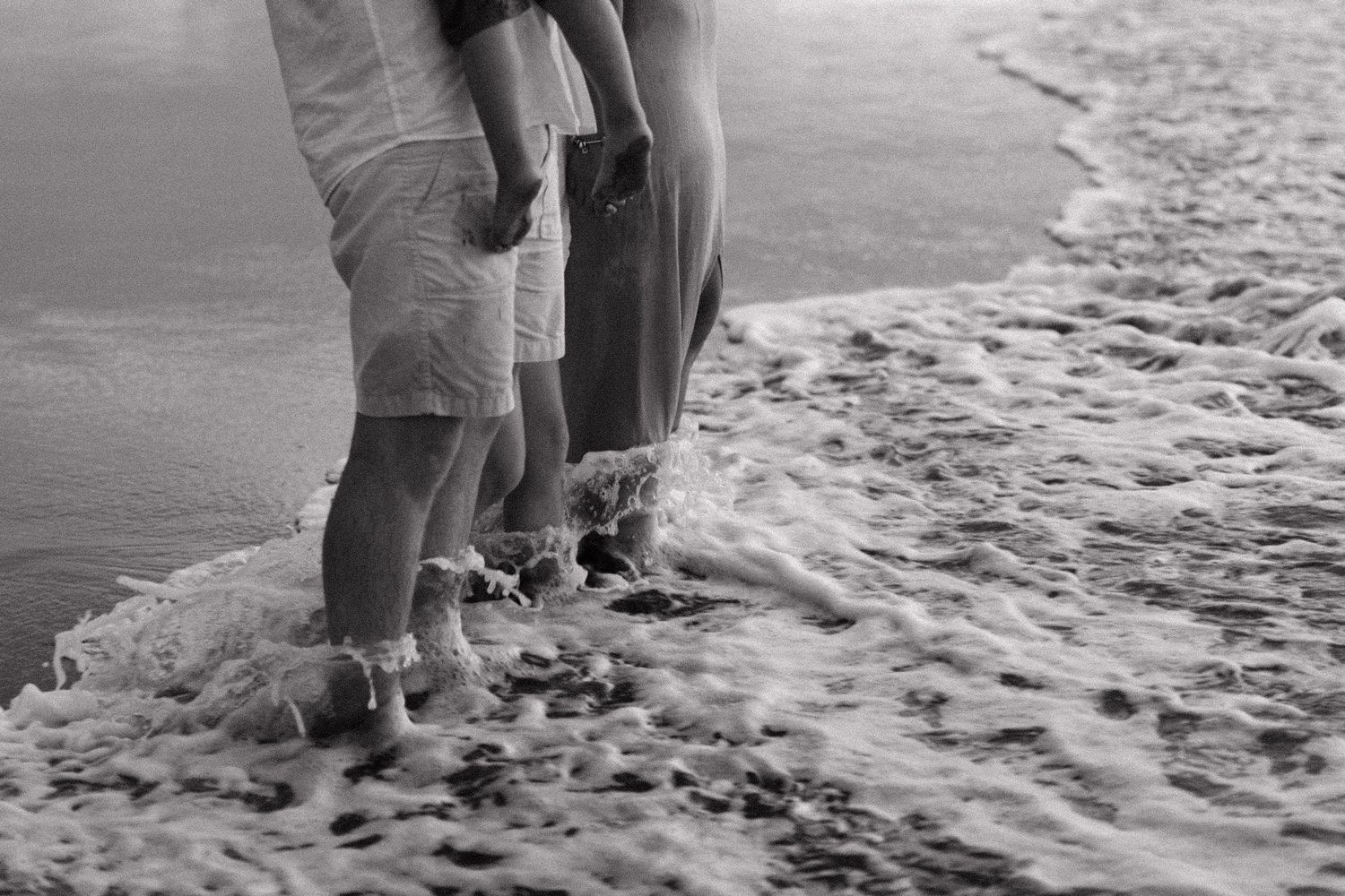 Person's legs walking barefoot along the sandy beach leaving footprints in shallow ocean water at sunset.