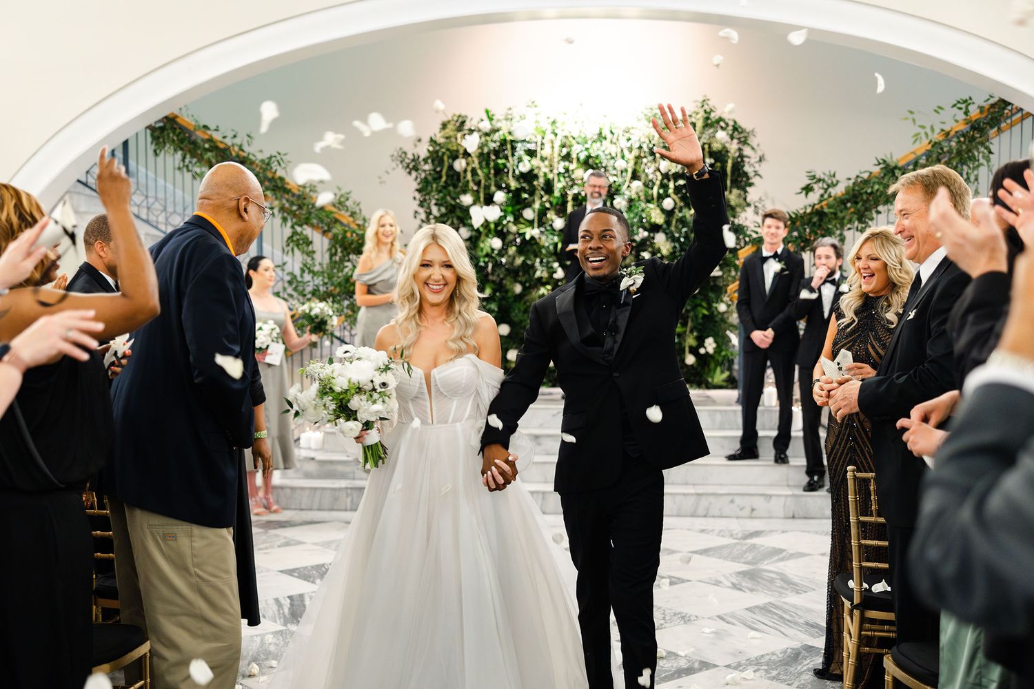 A newlywed couple walks down the aisle together as wedding guests cheer during a ceremony with floral decorations.