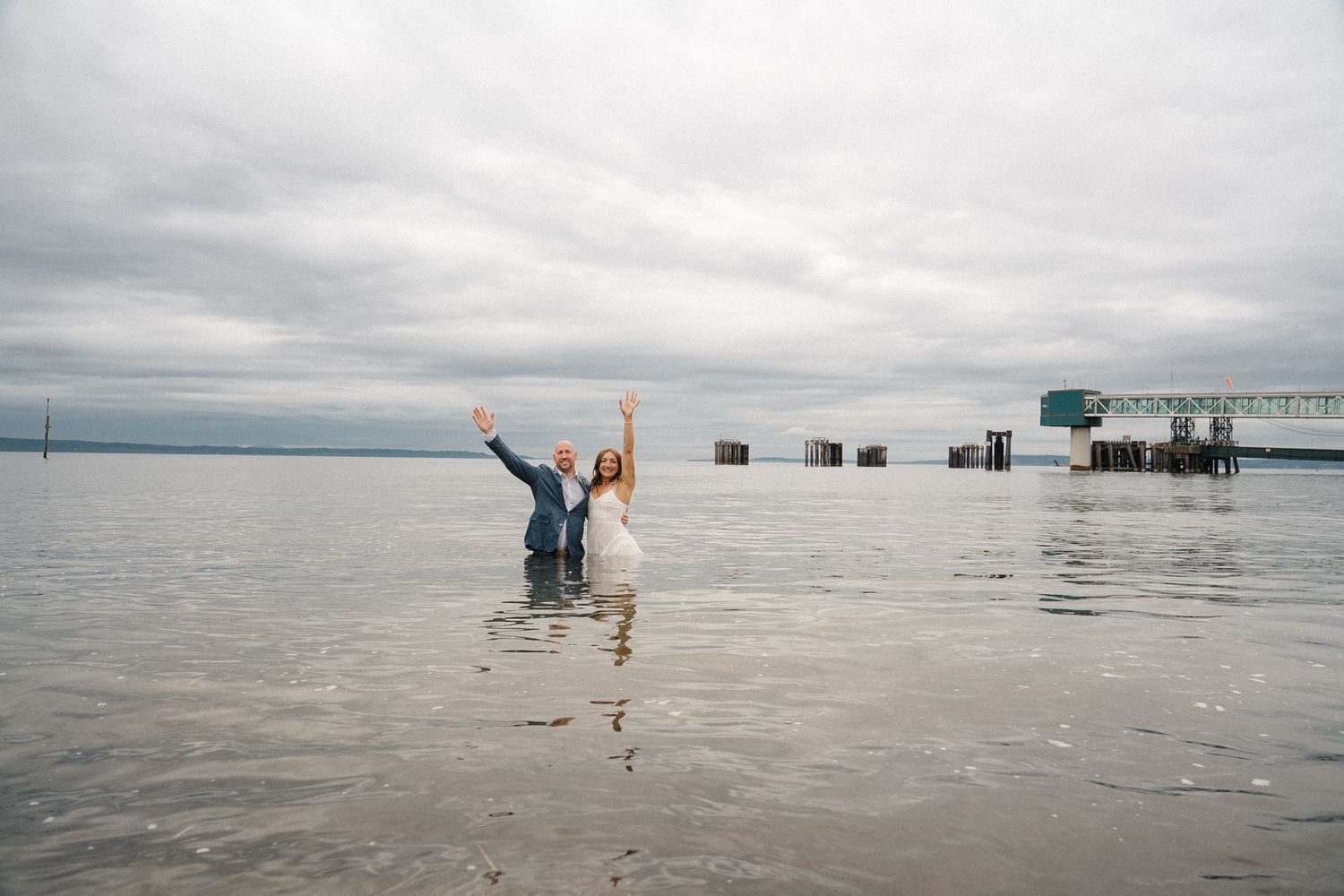 Two people wade playfully in shallow water with raised arms under cloudy skies at a coastal beach.