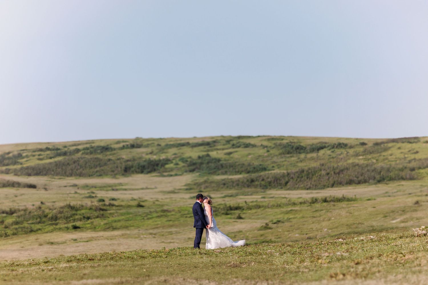 A couple embraces while standing on a rolling green hillside under soft blue skies.