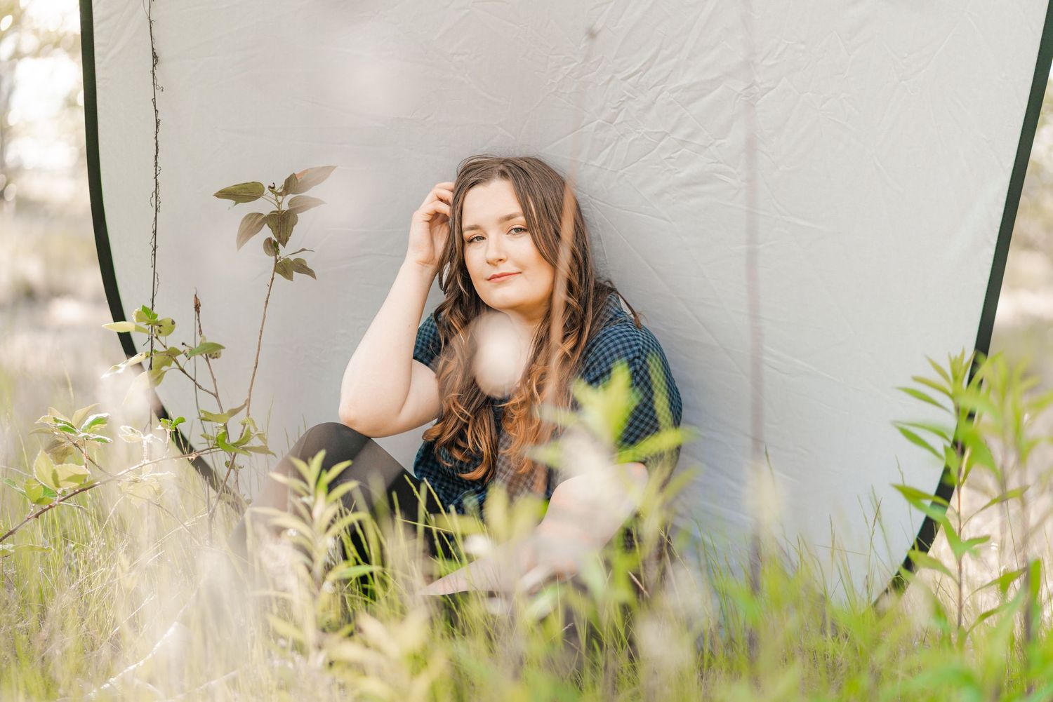 Young woman with long wavy hair sits against a neutral pop-up backdrop outdoors, surrounded by soft greenery and natural light.