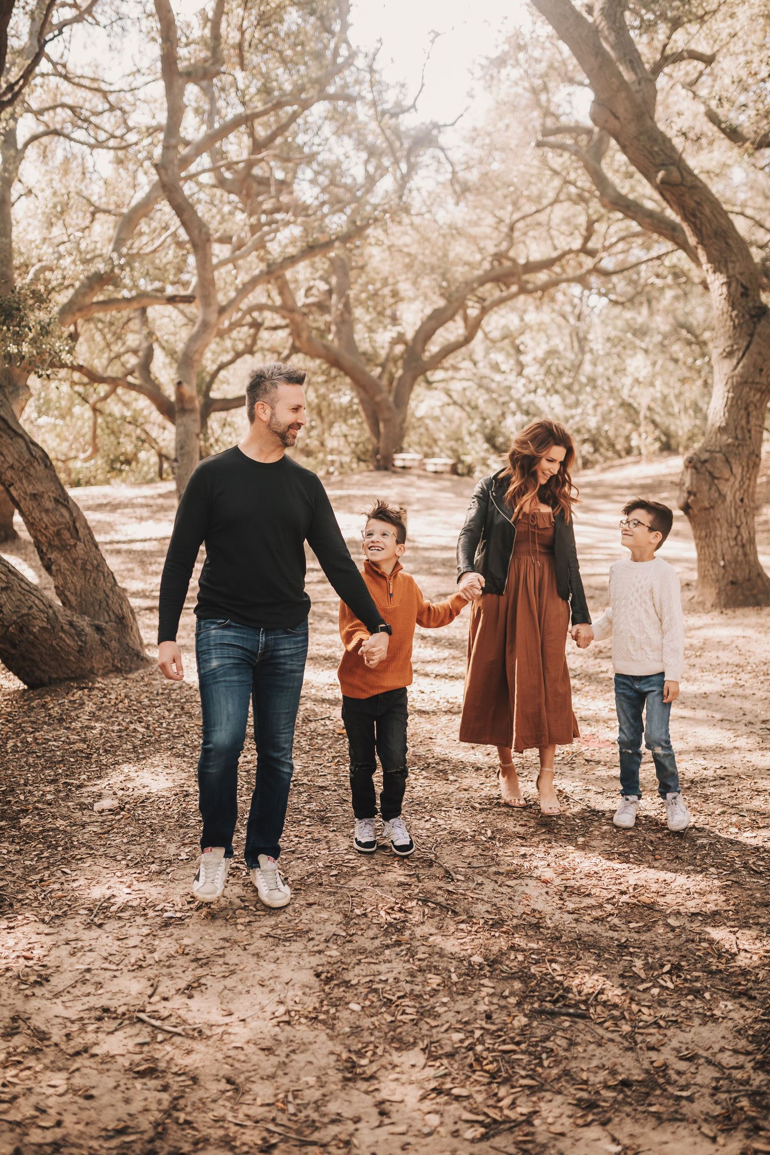 A family walks together holding hands through a wooded area with autumn leaves on the ground in warm muted tones.