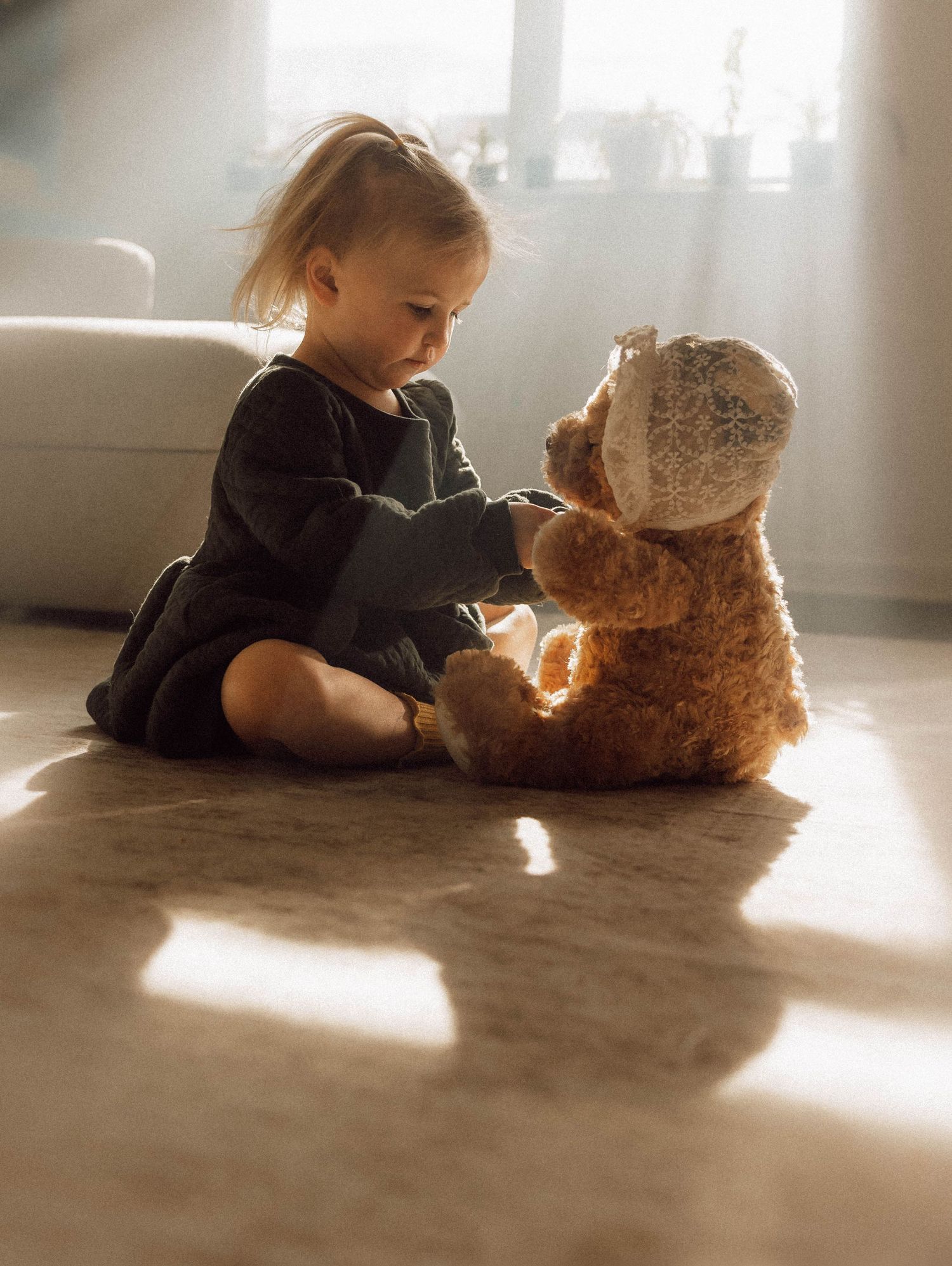 A small child in dark clothing lies on a carpeted floor beside a large teddy bear in warm sunlight streaming through a window.