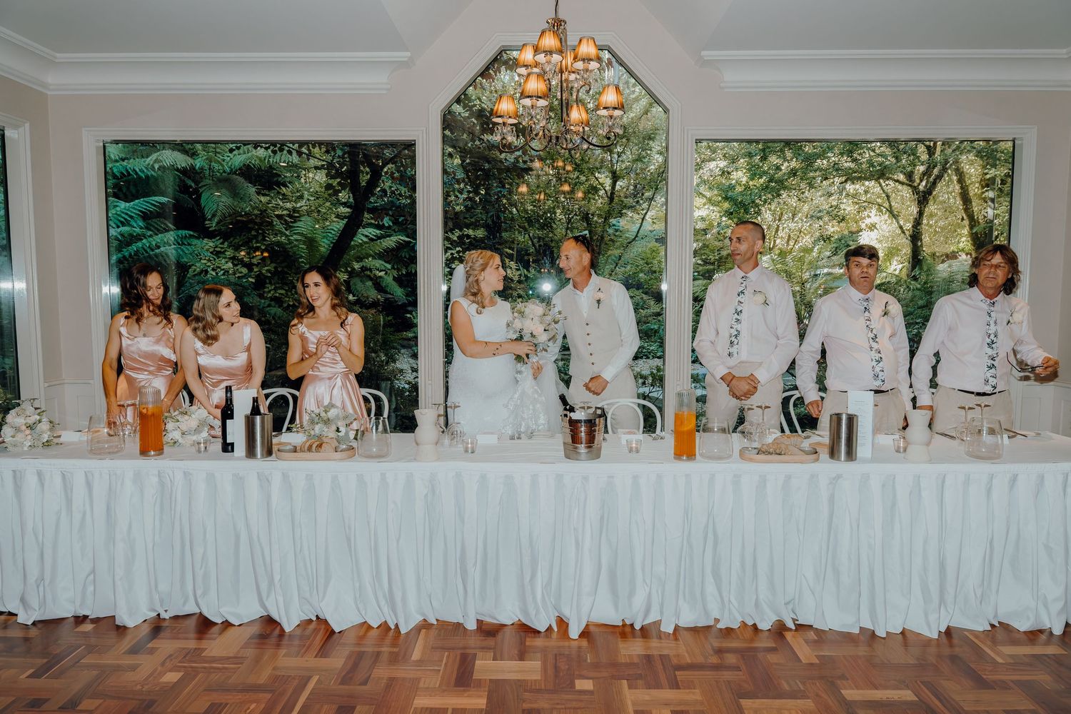 A group in white dresses and formal attire poses outdoors against a rustic wooden backdrop during a reception setup.