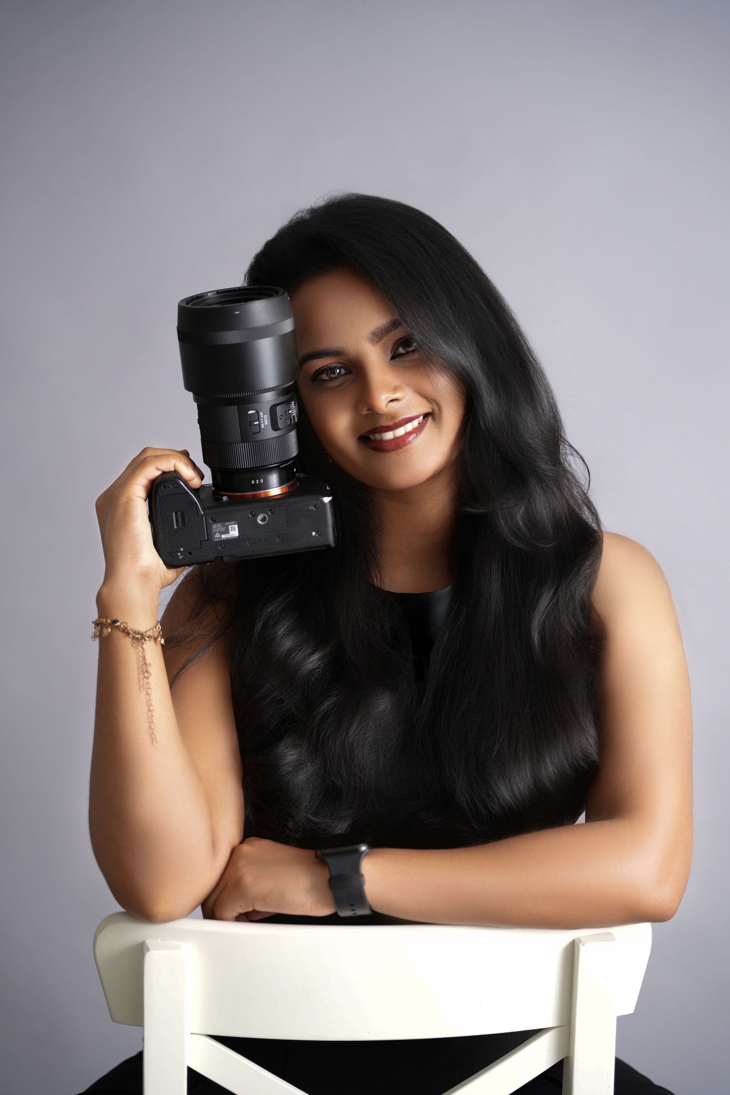 A photographer in a white top poses playfully with a professional camera lens against a light gray backdrop.