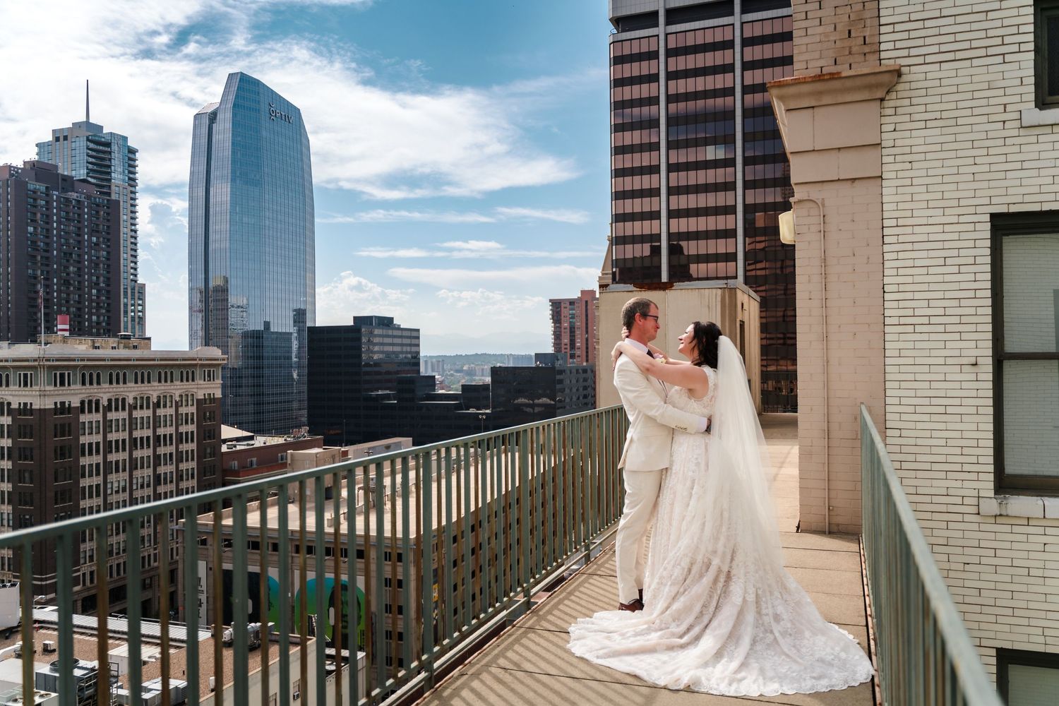 Couple in white wedding attire share intimate moment on urban rooftop terrace with downtown skyscrapers in background.