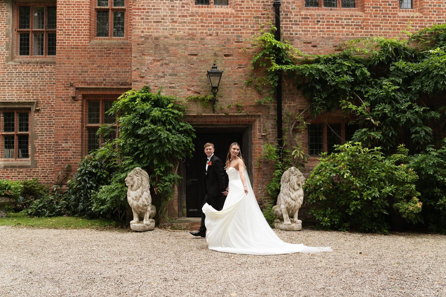 A bride in a white wedding gown poses by a historic brick building with garden statues and landscaping.