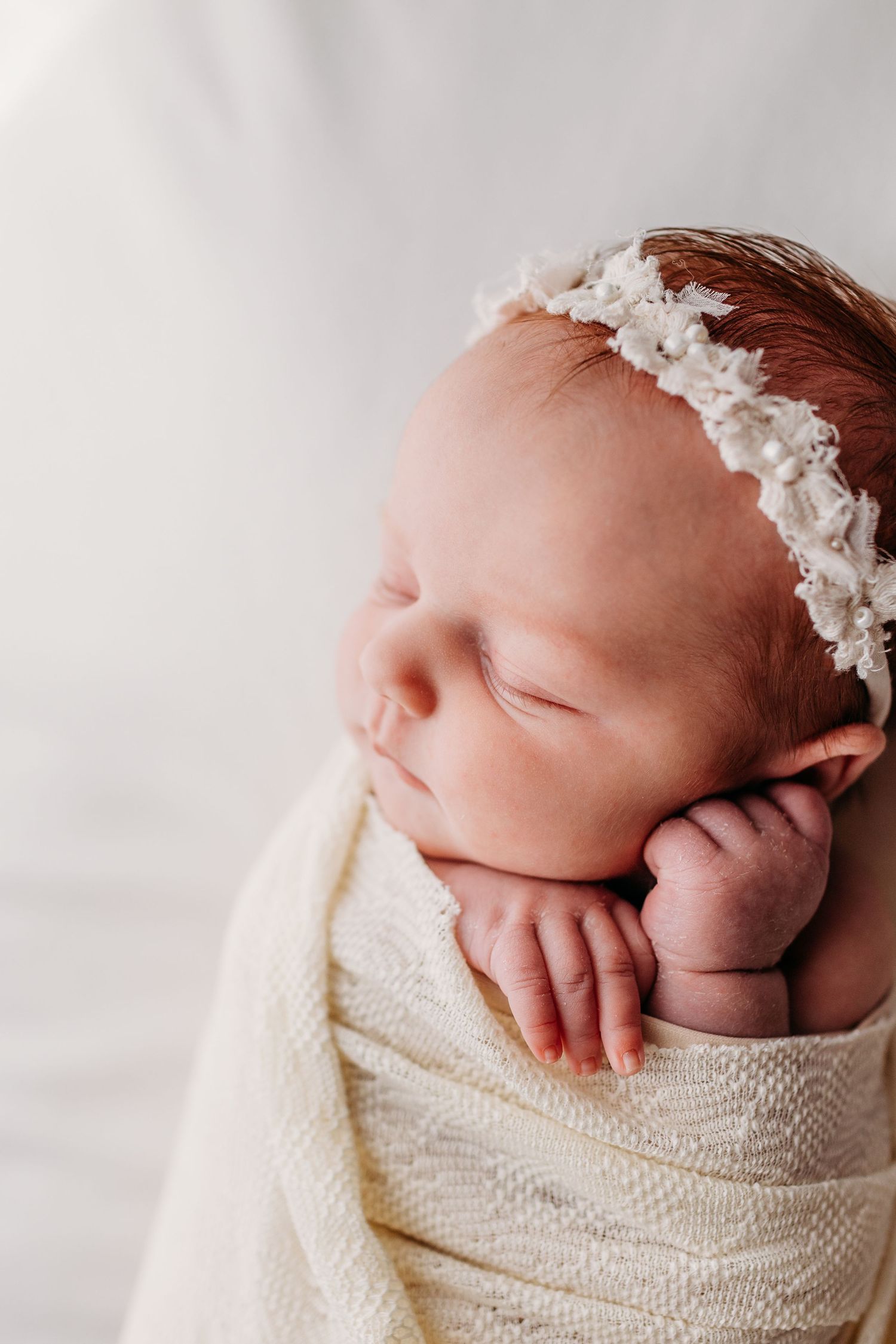 A sleeping newborn baby wrapped in a cream knit blanket wearing a delicate white floral headband.