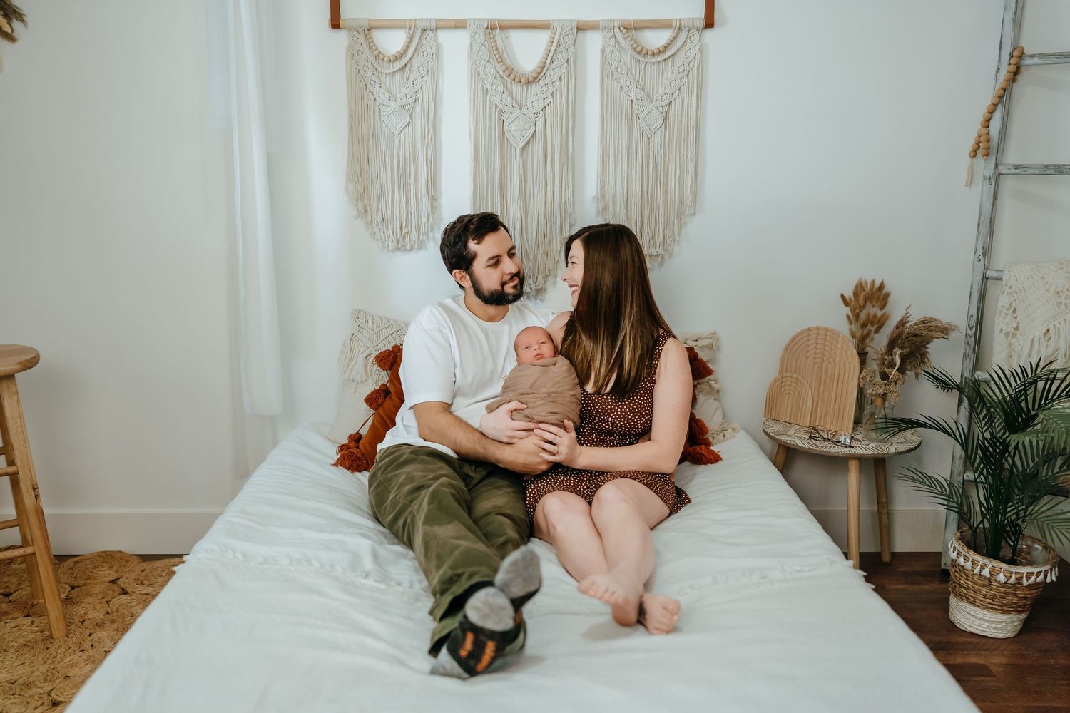 Intimate couple sits together on white bed near macrame wall hanging and potted plants.