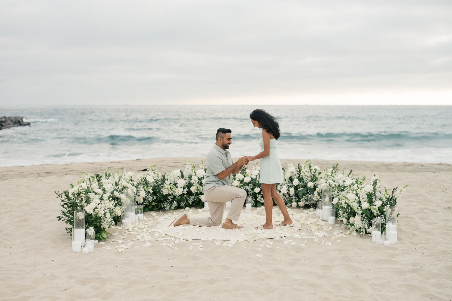 A romantic beach proposal scene with white floral arrangements decorating the sandy shoreline as waves roll in the background.