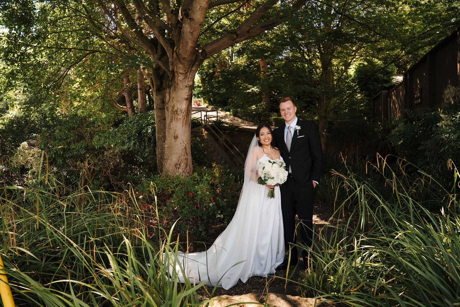 A couple in formal wedding attire stands beneath a large tree in a lush garden setting.