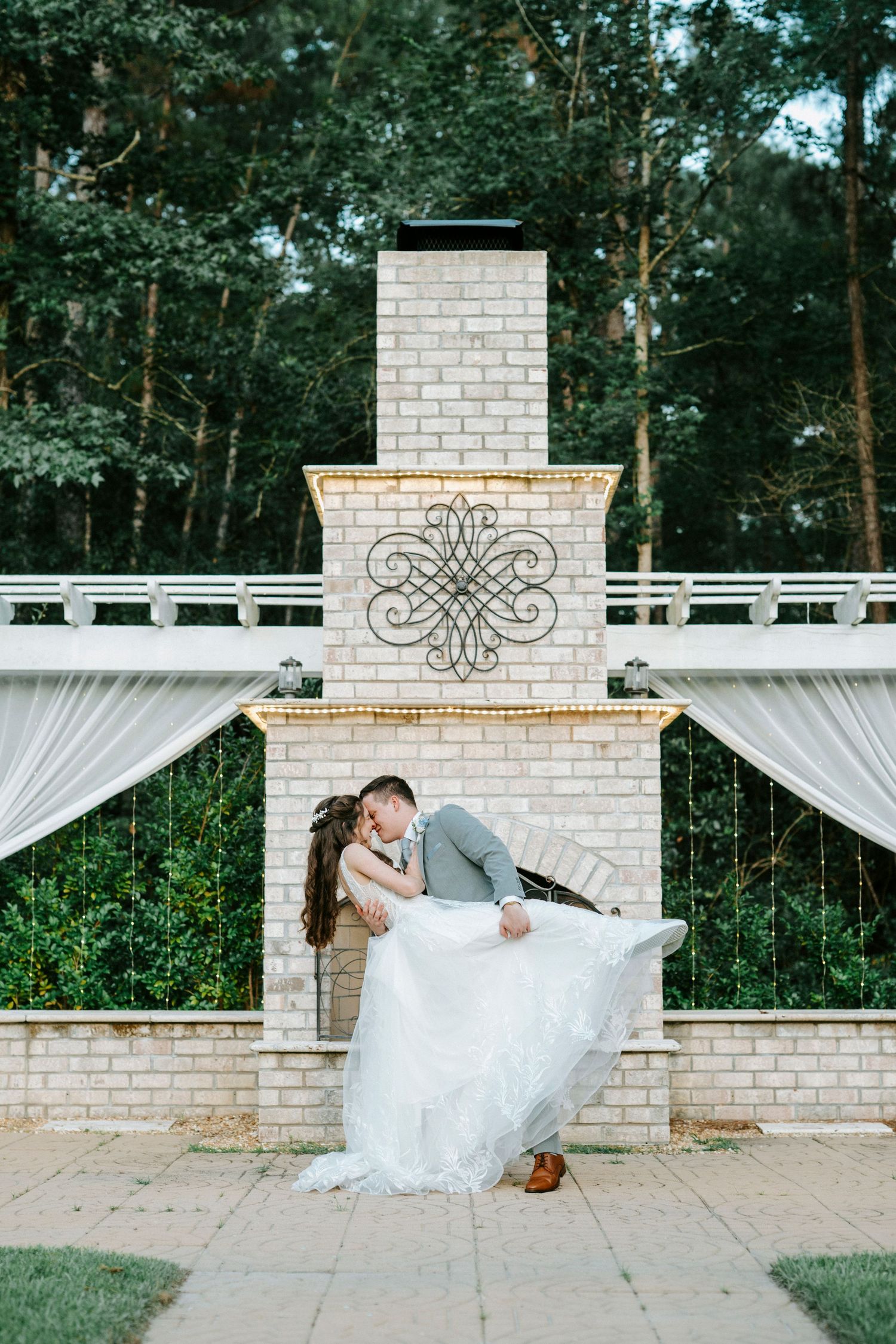 Wedding couple dips and kisses by an elegant white brick fireplace with decorative iron scroll details and flowing drapes.