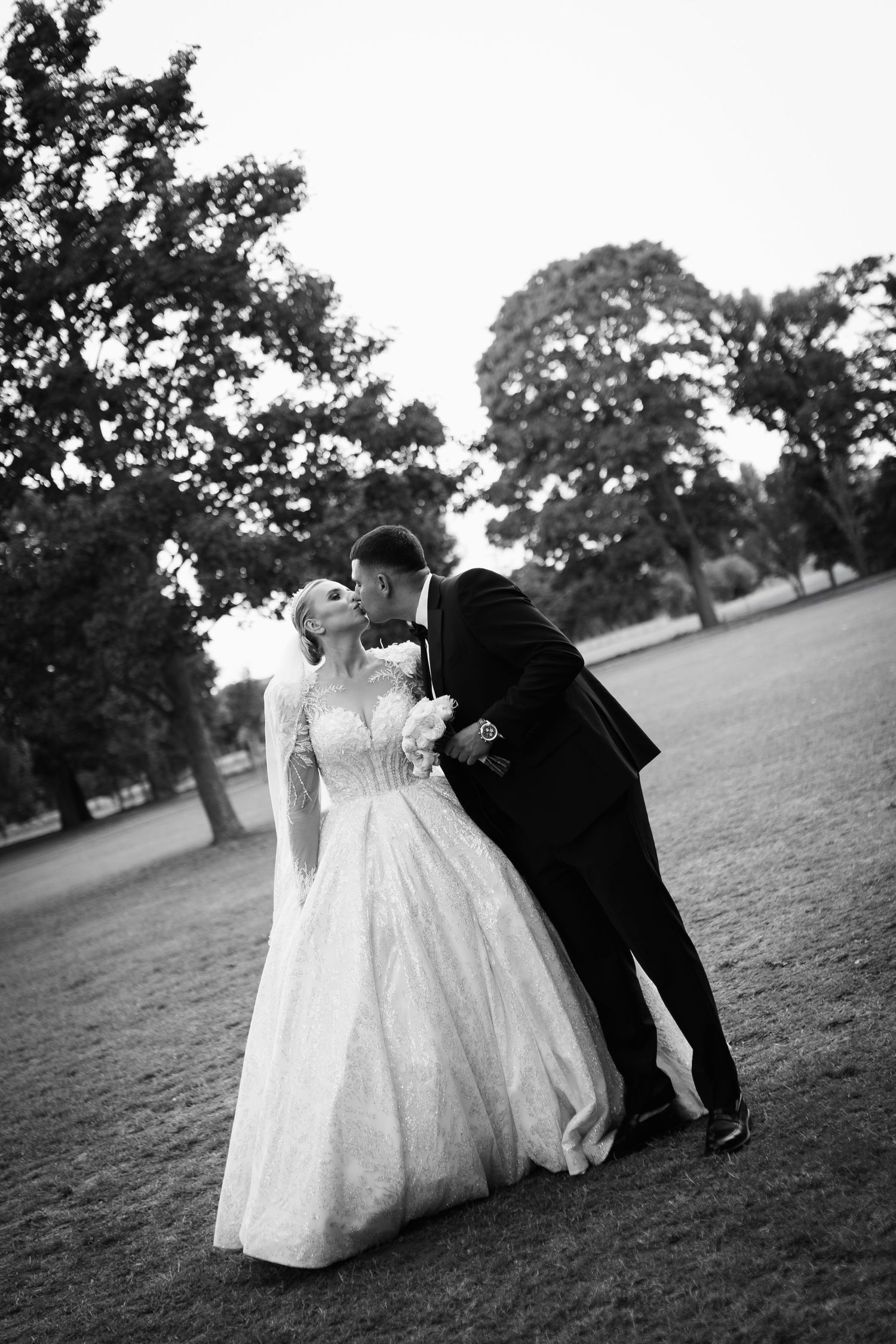 A couple share a romantic kiss on their wedding day against a backdrop of trees.