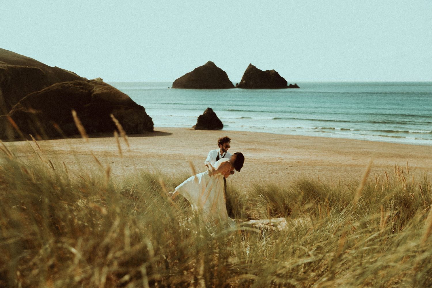 A scenic coastal landscape with tall grass swaying in the breeze against a turquoise ocean and rocky cliffs.