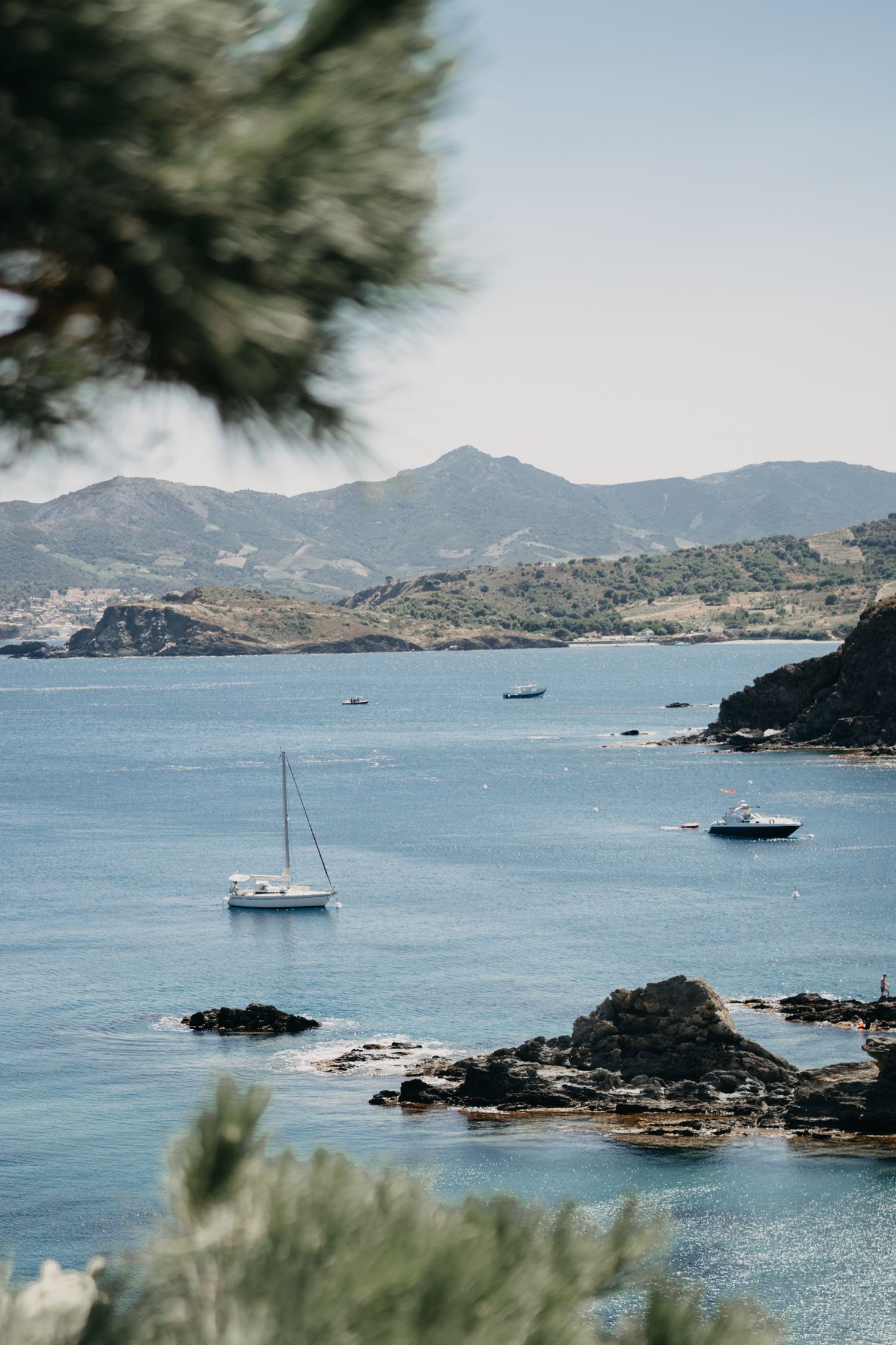 Aerial view of a small boat anchored in crystal clear turquoise waters near rocky Mediterranean coastline.