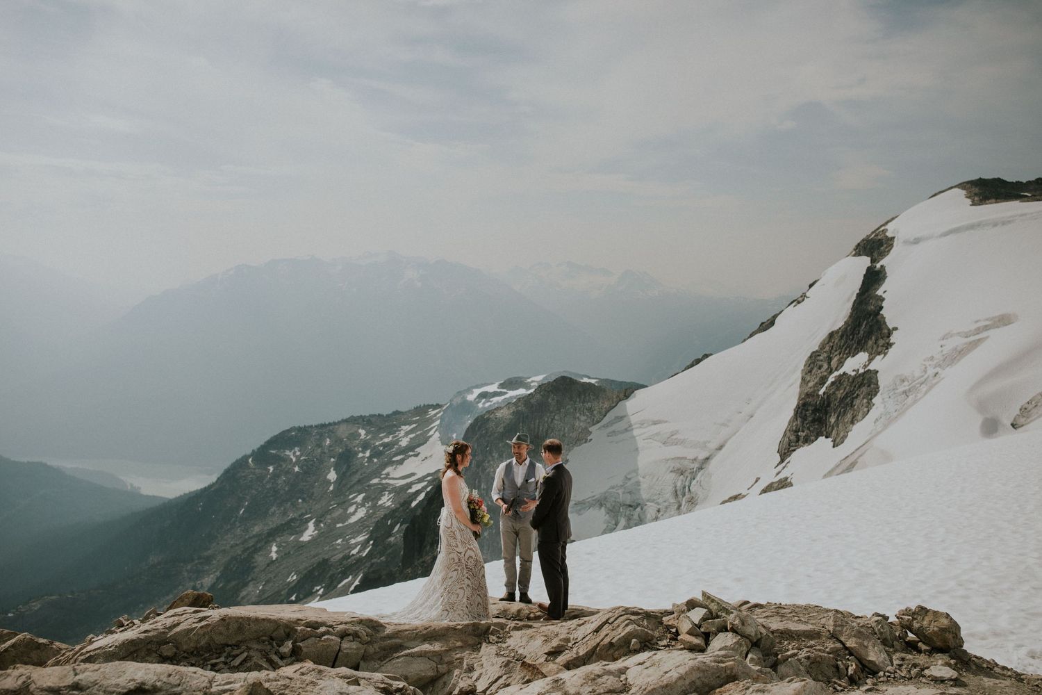 Couple stands on snowy mountain peak overlooking vast winter landscape during adventure engagement photoshoot.
