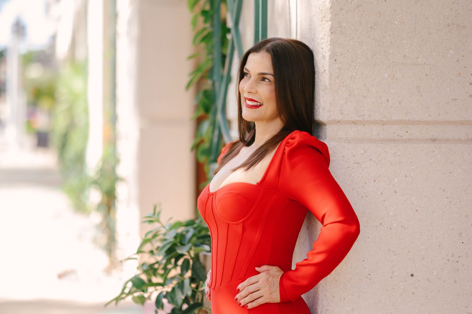 Woman in vibrant red dress poses confidently against light colored wall with green plants in background.
