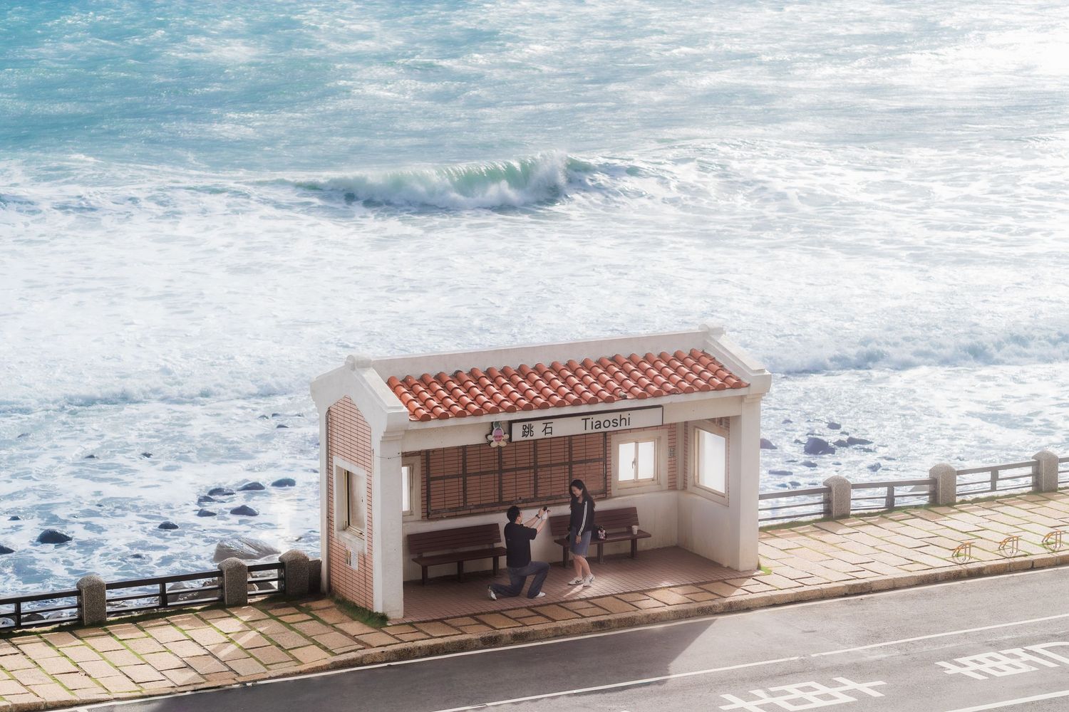 A quaint bus shelter with a tiled roof sits along a coastal road with crashing ocean waves in the background.
