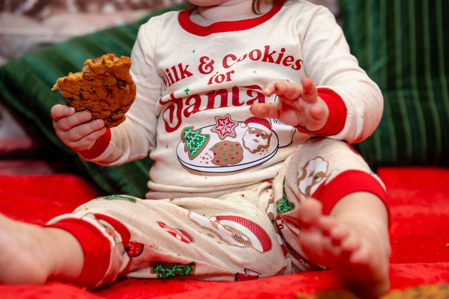 A baby wearing a white holiday-themed milk and cookies sweater sits on a red blanket.