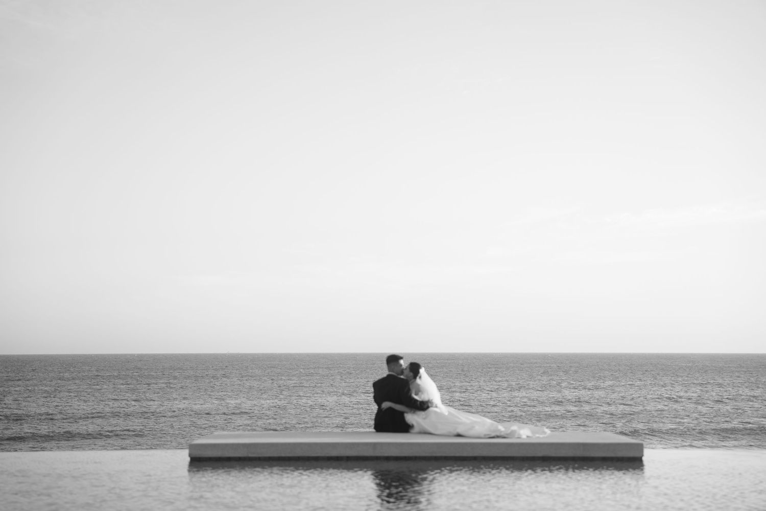 Black and white photo of bride and groom embracing on infinity pool edge overlooking calm ocean.
