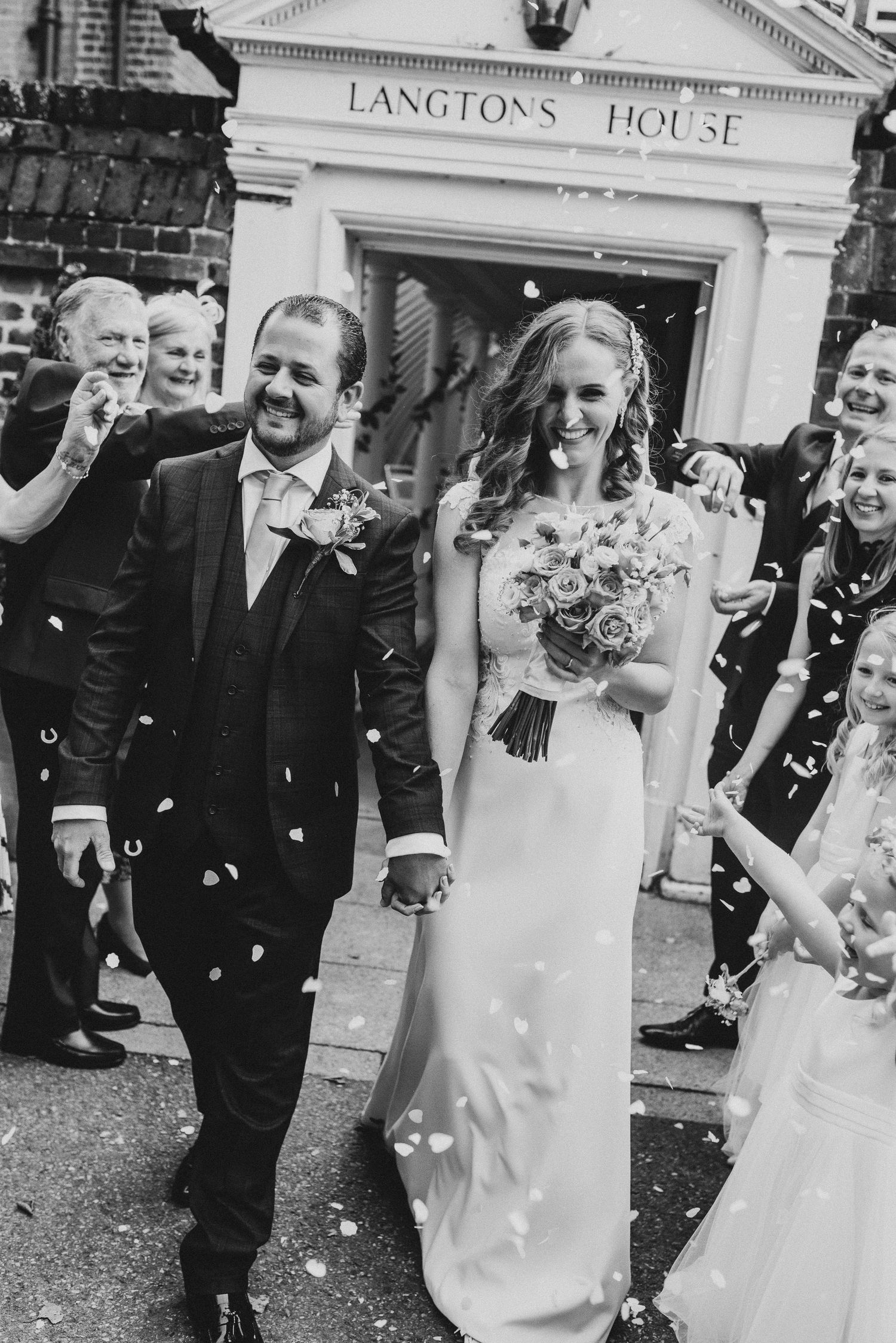 Black and white photo of a bride and groom walking out of Langtons House Registry Office, surrounded by guests throwing confetti.