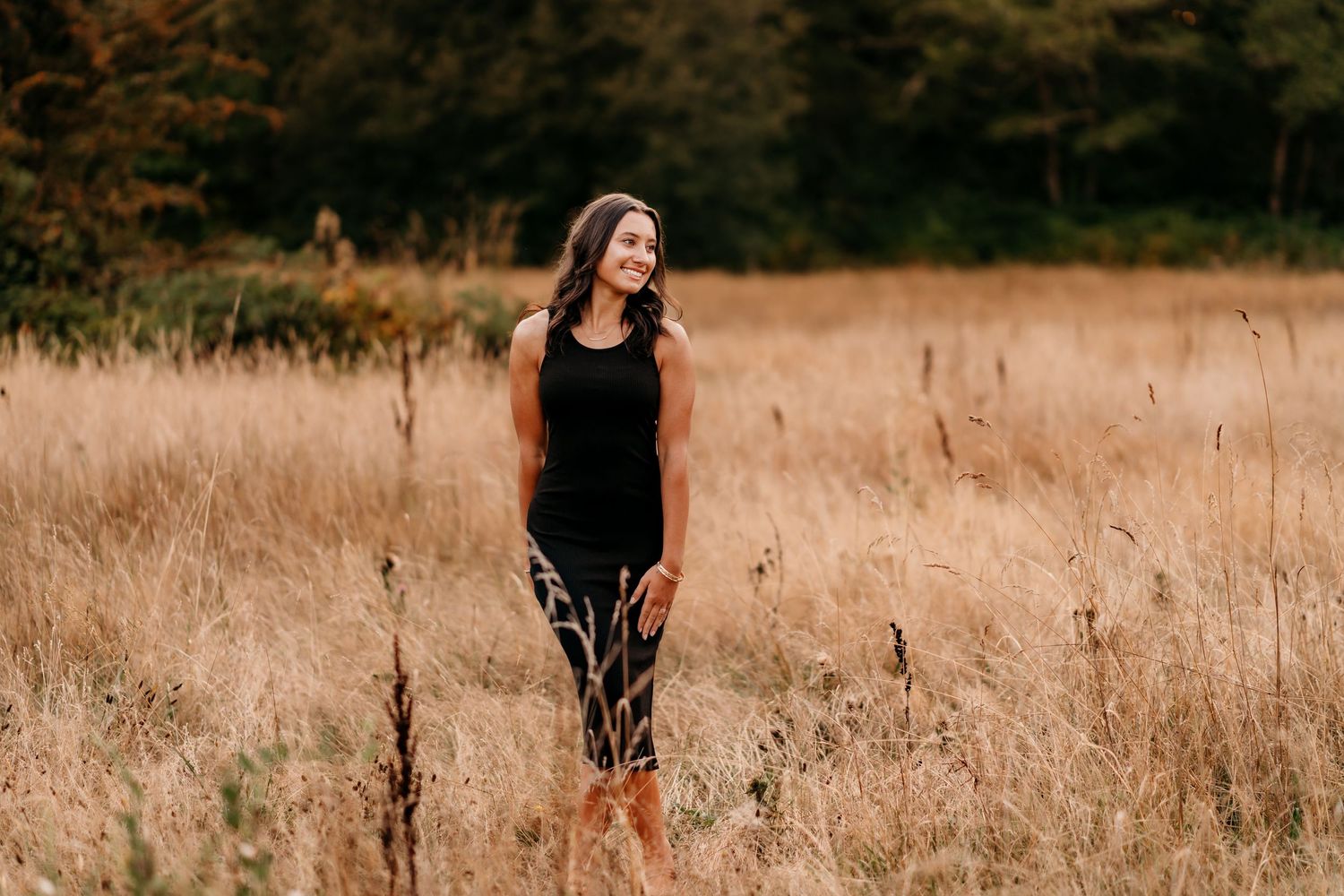 High school senior in a black dress poses gracefully in a golden field during sunset during senior photo session in Puyallup.