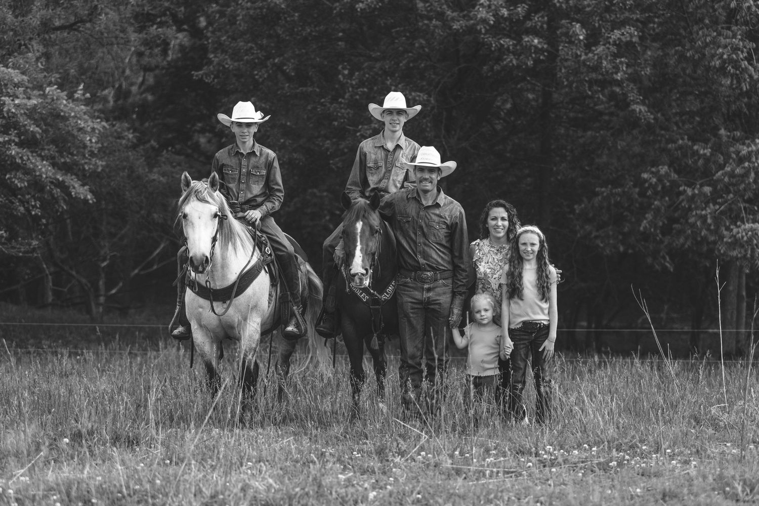 Black and white photograph showing two cowboys on horseback with other people standing in a grassy meadow.