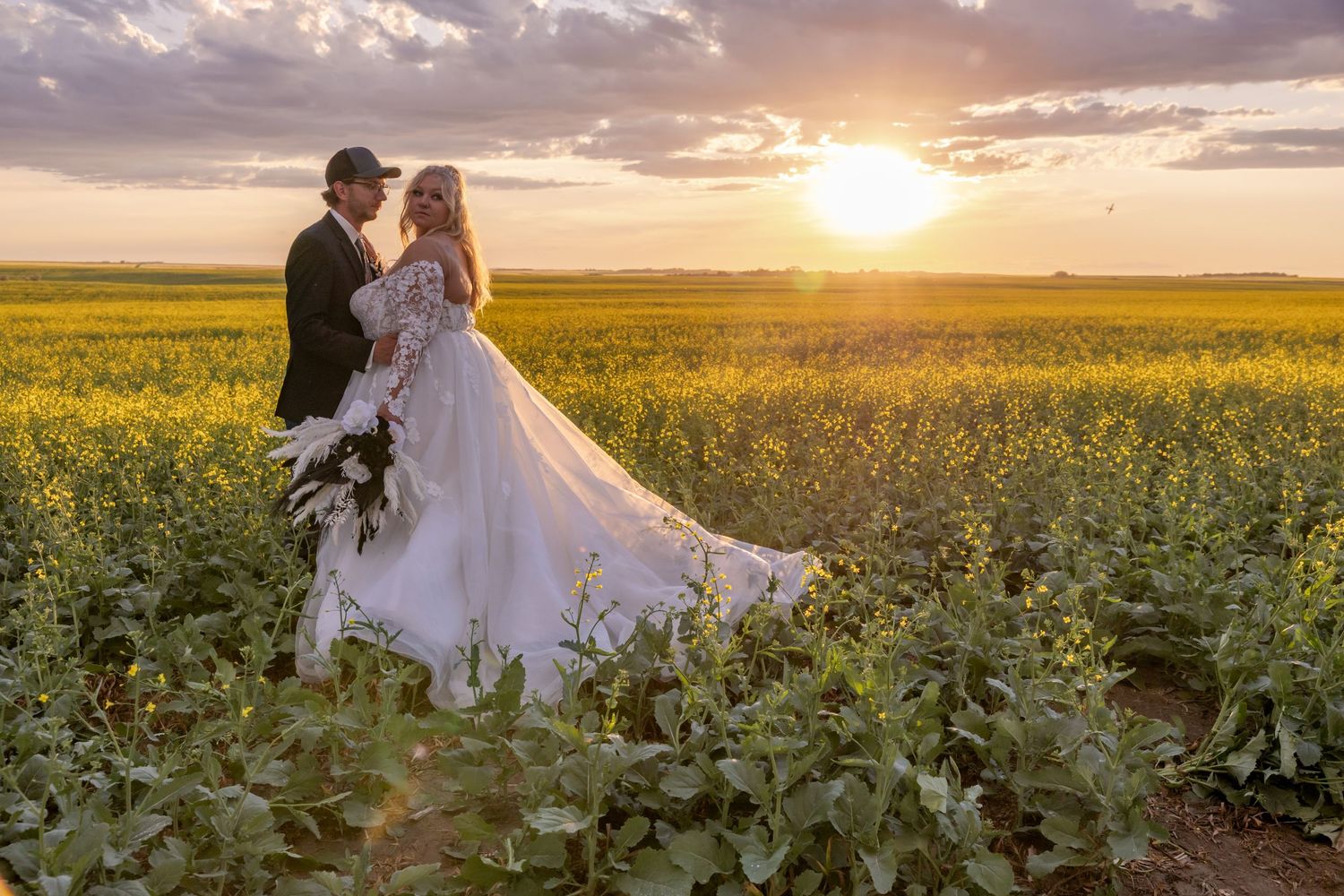 A romantic wedding couple stands in a beautiful yellow canola field at sunset with dramatic clouds in the sky. Garrison, ND