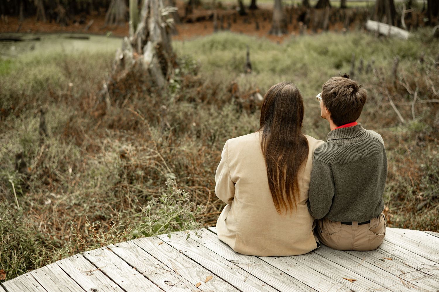 A couple sits together on a wooden bench in a rustic outdoor setting surrounded by autumn foliage.