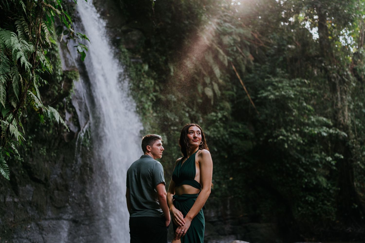 A couple poses against the backdrop of a scenic waterfall in a tropical forest setting.